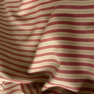 May include: A woven fabric featuring a repeating pattern of wide red stripes alternating with narrower off-white stripes. The material displays a visible textile weave with soft folds and drapes that create subtle shadows across the surface texture.
