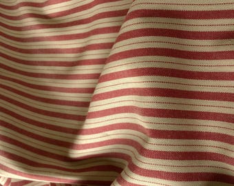 May include: A woven fabric featuring a repeating pattern of wide red stripes alternating with narrower off-white stripes. The material displays a visible textile weave with soft folds and drapes that create subtle shadows across the surface texture.