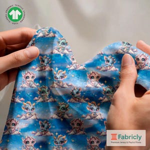 May include: Blue fabric featuring a repeating pattern of cartoon dragons on clouds. The dragons are coloured blue, pink, and green. The fabric is held up by hands, and the GOTS logo is visible in the upper left corner.