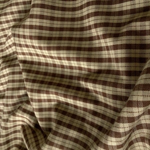 May include: A piece of fabric featuring a woven plaid pattern in shades of dark brown and off-white. The textile displays a consistent grid design with intersecting horizontal and vertical lines, creating a textured appearance across the folded surface.