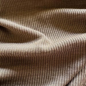 Peut inclure: Gros plan sur un tissu en maille côtelée texturé de couleur marron neutre et chaleureuse. Le tissu a un aspect doux et drapé avec des côtes verticales, créant un motif subtil. L'éclairage met en valeur la texture et les plis du matériau.