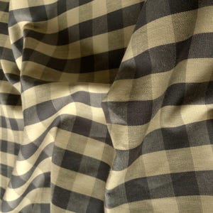 Peut inclure: Un tissu présentant un motif à carreaux vichy classique avec des carrés alternés gris anthracite foncé et beige clair. Le matériau est présenté avec des plis et des drapés doux, soulignant la texture tissée et le motif en grille régulier.