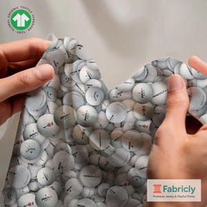Könnte beinhalten: Stoff mit Golfballmuster. Der Stoff ist weiß mit einem sich wiederholenden Muster aus Golfbällen. Der Stoff wird von zwei Händen gehalten. Der Stoff hat das "GOTS"-Logo und das Fabricly-Logo.