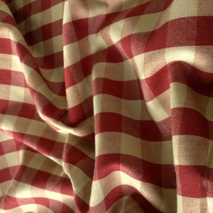 Op de afbeelding: Een stof met een klassiek rood en gebroken wit gingham ruitpatroon. Het materiaal is gedrapeerd om de textuur en zachte plooien te tonen, waarbij het consistente vierkante rasterontwerp over het gehele textieloppervlak wordt benadrukt.