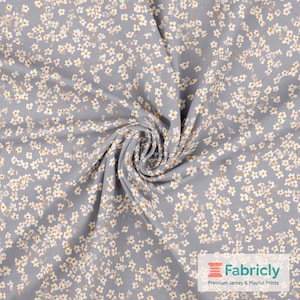 Könnte beinhalten: Nahaufnahme eines Stoffes mit floralem Muster. Der Stoff ist hellgrau und mit kleinen weißen Blumen mit gelben Zentren bedeckt. Der Stoff ist in einer Spirale zusammengefasst, wodurch die Textur und das Muster sichtbar werden.