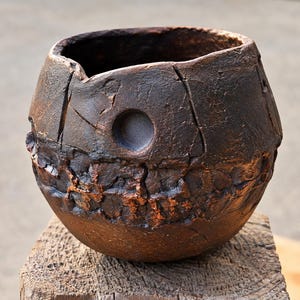 May include: A dark brown, spherical ceramic bowl with a heavily textured, cracked surface. The exterior features a prominent horizontal band of deep, rugged fissures and a single circular indentation near the rim, showcasing a rustic, weathered finish.