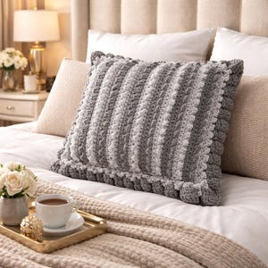 May include: A gray and white striped crochet pillow with a ruffled edge. The pillow is rectangular and sits on a bed with white sheets and a beige blanket. A gold tray with a cup of coffee and flowers is in the foreground.
