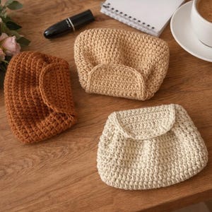 Puede incluir: Tres monederos de ganchillo hechos a mano en tonos marrón y beige se exhiben sobre una superficie de madera. Los monederos tienen un aspecto texturizado y diseños variados, uno de ellos con cierre de solapa. También se ven una taza de café, flores y un bolígrafo.