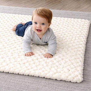 Puede incluir: Un bebé sonriente con una camisa gris de manga larga y vaqueros azules está acostado sobre una manta blanca texturizada. La manta está sobre una alfombra gris. El bebé mira a la cámara.