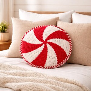 Peut inclure: Un coussin rond en crochet rouge et blanc, avec un motif en spirale rappelant une canne en sucre d'orge. Le coussin, d'environ 35 cm de diam&egrave;tre, est bord&eacute; d'une couture rouge et blanche. Il est pos&eacute; sur un lit avec des coussins neutres.