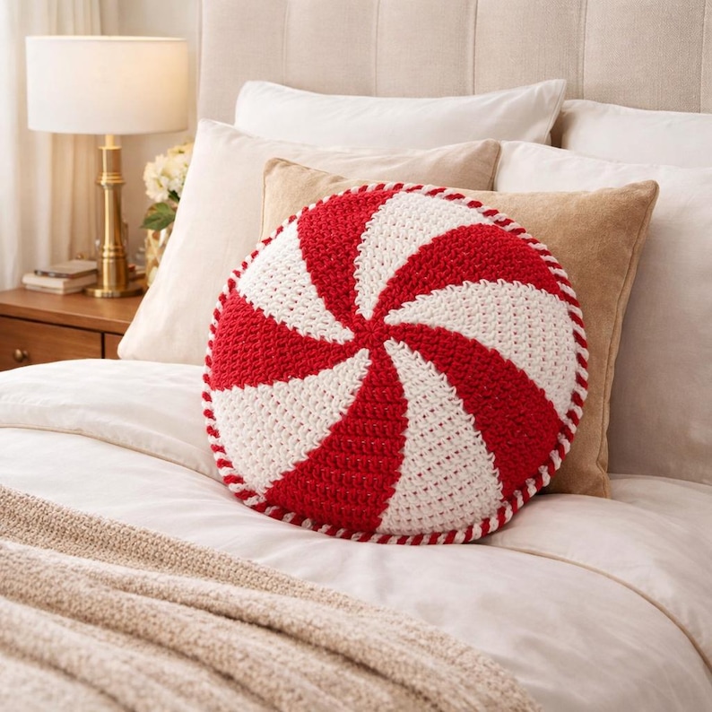 Peut inclure: Un coussin rond en crochet &agrave; rayures rouges et blanches, rappelant un bonbon &agrave; la menthe poivr&eacute;e. Le coussin est pos&eacute; sur un lit avec des draps blancs et une couverture beige. D&eacute;coration d'int&eacute;rieur festive.