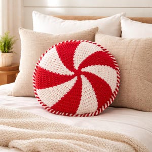 Peut inclure: Un coussin rond en crochet rouge et blanc avec un motif en spirale rappelant une canne en sucre d'orge. Le coussin a une bordure rouge et est pos&eacute; sur un lit avec des oreillers et une couverture de couleurs neutres. Le coussin mesure environ 35 cm de diam&egrave;tre.