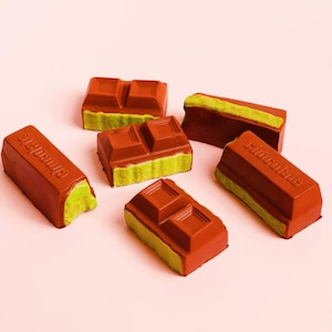 May include: A collection of reddish-brown chocolate pieces with a green base, arranged on a pale pink surface. Some pieces are embossed with the word "Chocolate". The chocolate bars are rectangular and have a textured surface. The image shows a close-up of the chocolate.