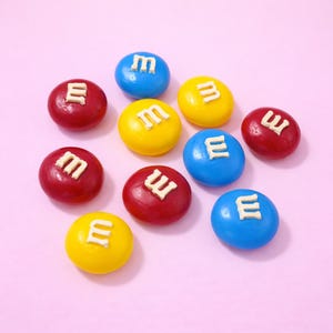 May include: A selection of colourful sweets in red, yellow, and blue, each with a white "E" or "M" printed on them. The sweets are scattered on a light pink background.