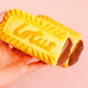 Puede incluir: Dos pasteles rectangulares amarillos con relleno marrón se sostienen en una mano sobre un fondo rosa. La palabra "Lotus" está escrita en la parte superior de los pasteles en amarillo-naranja.