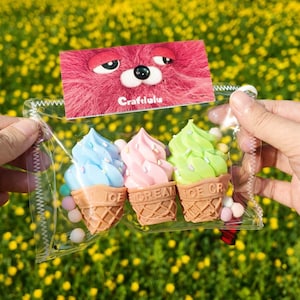 May include: A clear plastic bag containing three ice cream cone-shaped ornaments with blue, pink, and green soft serve. The cones have the word "ICE CREAM" on them. A pink card with a cartoon bear face is attached to the bag.