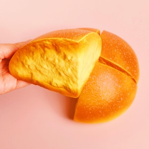 Puede incluir: Un pan cortado en cuatro trozos, con una corteza dorada y un interior suave y texturizado. Una mano sostiene un trozo, los otros tres están sobre un fondo rosa. El pan parece fresco y apetitoso.