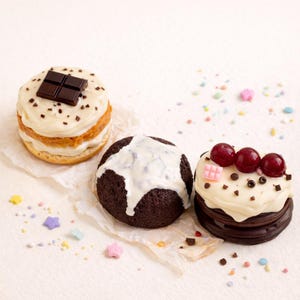 Puede incluir: Tres pastelitos con diferentes coberturas están colocados sobre una superficie blanca. Uno tiene una barra de chocolate encima, otro tiene glaseado blanco y el tercero tiene cerezas y chispas de chocolate. Hay chispas de colores esparcidas.