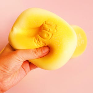 Puede incluir: Un juguete blando amarillo con forma de vaca está siendo apretado por una mano. El juguete tiene un diseño de cara de vaca y es un artículo suave para aliviar el estrés. El fondo es de color rosa liso.