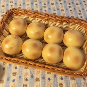 May include: A rectangular woven wicker basket containing eight round, smooth, light yellow buns with subtle brown toasted spots. The basket features a braided rim and a light brown finish, holding the uniform, spherical items in a neat arrangement.