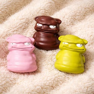 Puede incluir: Tres figuras de dibujos animados coloridas con expresiones gruñonas. Una es rosa, otra marrón y otra amarilla. Cada una tiene ojos blancos y una boca ancha y curvada. Las figuras están sobre una superficie suave y blanca.