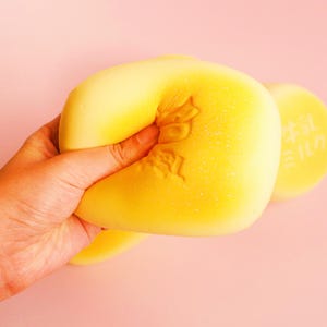 May include: A hand squeezes a yellow, squishy toy with a textured surface. The toy has an embossed design and is partially compressed. Another yellow toy with Japanese text is visible in the background against a pink backdrop.