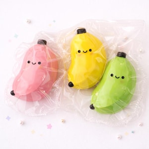 May include: Three colorful banana-shaped figurines in clear plastic packaging. The bananas are pink, yellow, and green, each with a black stem and smiling face. The background is white with scattered decorative elements.