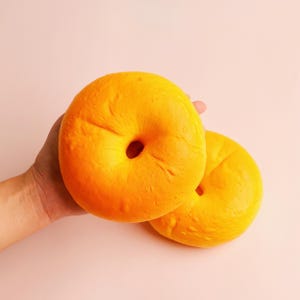 May include: Two round, golden-yellow bagels with a smooth, slightly textured surface and a central hole. The bagels have a uniform, vibrant color and a rounded, doughy appearance, showcasing a classic circular shape with a soft, baked-like finish.