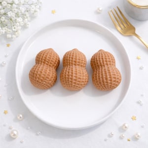 Puede incluir: Tres objetos con forma de cacahuete y superficie texturizada están dispuestos sobre un plato blanco. Los objetos son de color marrón claro y tienen un patrón detallado en forma de rejilla. El plato está sobre una superficie blanca, con elementos decorativos como perlas y un tenedor dorado.