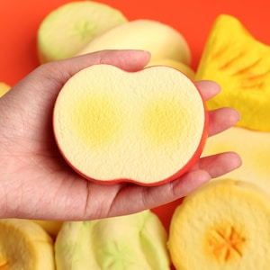 Puede incluir: Una mano sostiene una rodaja de manzana de espuma con borde rojo y centro amarillo. Otras rodajas de fruta de espuma están en el fondo sobre un fondo naranja. La rodaja de manzana tiene una apariencia realista.