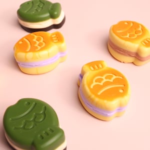 Puede incluir: Un surtido de coloridas golosinas con forma de pez. Las golosinas son en tonos verdes, amarillos y naranjas, con acentos morados y blancos. Las golosinas están dispuestas sobre una superficie rosa claro.