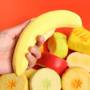 Puede incluir: Una mano sosteniendo un objeto amarillo con forma de plátano. Otros objetos coloridos con forma de fruta, incluyendo manzanas y piña, están esparcidos sobre un fondo rojo. El plátano es el punto focal, con una superficie texturizada.
