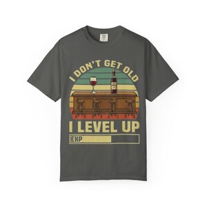 Puede incluir: Camiseta gris oscuro con un diseño gráfico que presenta una escena de bar con una copa y una botella de vino. El texto dice "I DON'T GET OLD I LEVEL UP EXP". El diseño tiene un estilo retro.