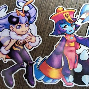May include: Two colorful stickers featuring anime-style characters. One is a bee-themed figure with purple hair and a fluffy collar. The other is a blue-skinned character in a pink and purple outfit, holding a spiky ball.