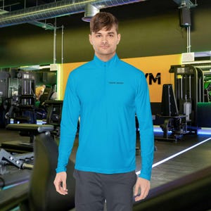 Op de afbeelding: Een turquoise sportshirt met lange mouwen en een kraag met een kwartrits. Het shirt heeft de tekst "SWEET BRAND" in het zwart op de borst. De persoon draagt een grijze sportbroek in een sportschool.