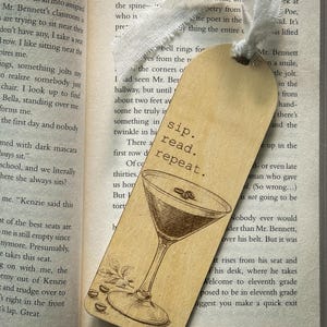 以下が含まれることがあります： 木製のブックマーク。マティーニのイラストと「sip. read. repeat.」の文字が黒色で描かれています。ブックマークは薄茶色の背景で、上部には白いリボンが付いています。開いた本の中に置かれています。