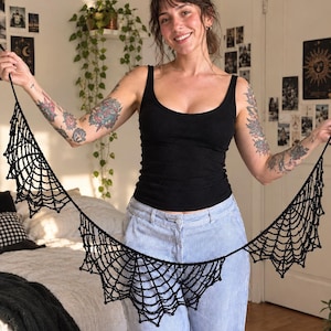 May include: A black crocheted garland featuring a series of interconnected spider web motifs. The decorative piece is constructed from dark yarn, forming a repeating pattern of web-like segments that are linked together by a thin, continuous cord.
