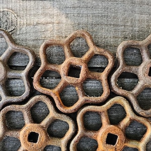 May include: Five circular metal valve handles with a rusted, weathered brown finish. Each handle features a five-spoke design with a square center opening. The handles are arranged in a cluster, showcasing their textured, oxidized surfaces.