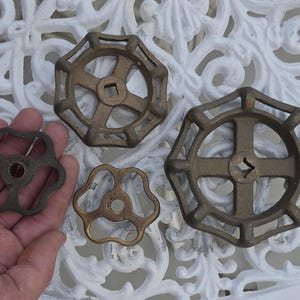 May include: Four metal valve handles in varying sizes and shapes. Two handles feature a circular, multi-spoke design with a square center opening, while the other two are smaller, three-lobed handles with a circular center hole, all in a bronze tone.