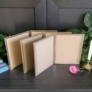 May include: Five gold picture frames with a light brown mat are arranged in a row on a black surface. The frames are all the same size and have a simple, elegant design.