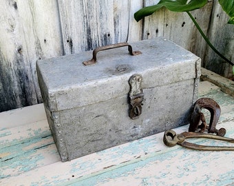 May include: A vintage metal toolbox with a hinged lid, a metal latch, and a metal handle. The toolbox is gray and has a worn, distressed look. It is sitting on a wooden surface with two rusty metal tools.