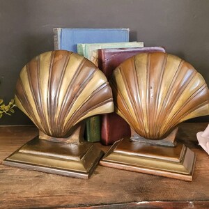 May include: A pair of brass bookends shaped like seashells. The bookends are a light gold color and have a detailed design. They are sitting on a wooden surface with a few books behind them.
