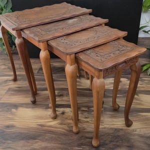 May include: A set of four graduated nesting tables crafted from medium-toned wood. Each table features a rectangular top with intricate carved floral borders and curved cabriole legs that terminate in simple, rounded feet.