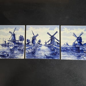 May include: Three square ceramic tiles featuring a blue and white transferware style landscape. Each tile depicts a Dutch scene with windmills, waterways, small boats, and rural buildings under a cloudy sky, rendered in detailed blue ink.