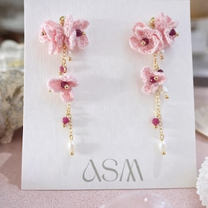 May include: A pair of pink floral earrings with gold-coloured chains and accents. The earrings feature crocheted flowers, small red beads, and dangling pearls. The earrings are displayed on a white card with the letters "ASM" in the bottom corner.