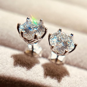 Può includere: Un paio di orecchini a lobo con diamanti scintillanti. Ogni orecchino presenta un grande diamante rotondo taglio brillante incastonato in una montatura argentata a griffe. I diamanti riflettono la luce, creando un'esposizione abbagliante.