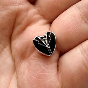 Könnte beinhalten: Eine herzförmige Anstecknadel mit einem schwarzen Smoking-Design. Die Anstecknadel zeigt einen schwarzen Smoking-Jackett mit weißem Hemd und Fliege. Die Anstecknadel hat einen silbernen Rand und drei kleine silberne Knöpfe.