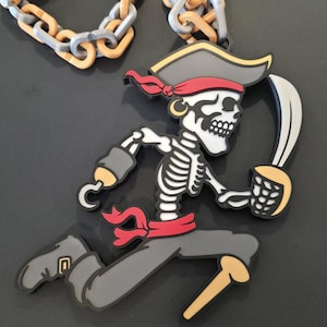 May include: A large pendant featuring a cartoon skeleton pirate wearing a hat, a red headband, and a red sash, holding a curved sword in one hand and a hook on the other arm. The skeleton has a peg leg and is attached to a thick, chunky plastic chain.