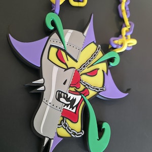 Op de afbeelding: Een kleurrijke hangerketting met een gespleten gezichtsontwerp. De ene kant heeft een geel en rood gezicht, de andere kant een grijs gezicht. De ketting heeft een ketting van paarse en gele schakels. De hanger heeft groene, paarse en zwarte accenten.