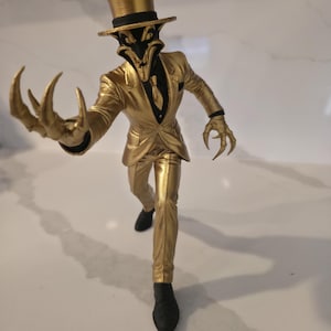 Pode incluir: Estatueta dourada de uma criatura de fato e cartola. A figura tem garras afiadas e uma expressão ameaçadora. O fato é dourado brilhante e a figura está em movimento.